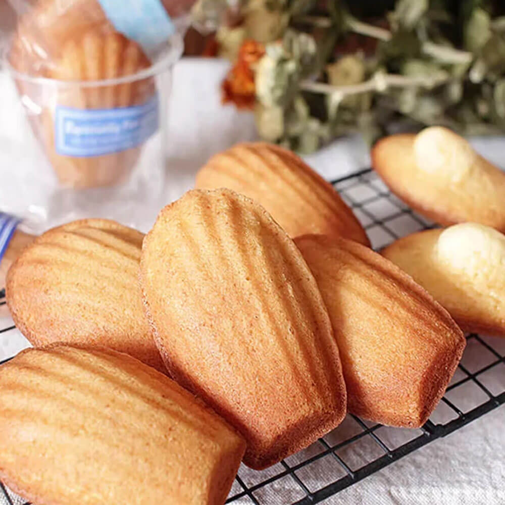 A golden-brown madeleines baked to perfection, displayed on a cooling rack. The madeleines have a classic shell-like pattern on their surface, achieved using a carbon steel baking pan. The surrounding setup includes a soft focus of dried flowers and a packaged container of similar treats.