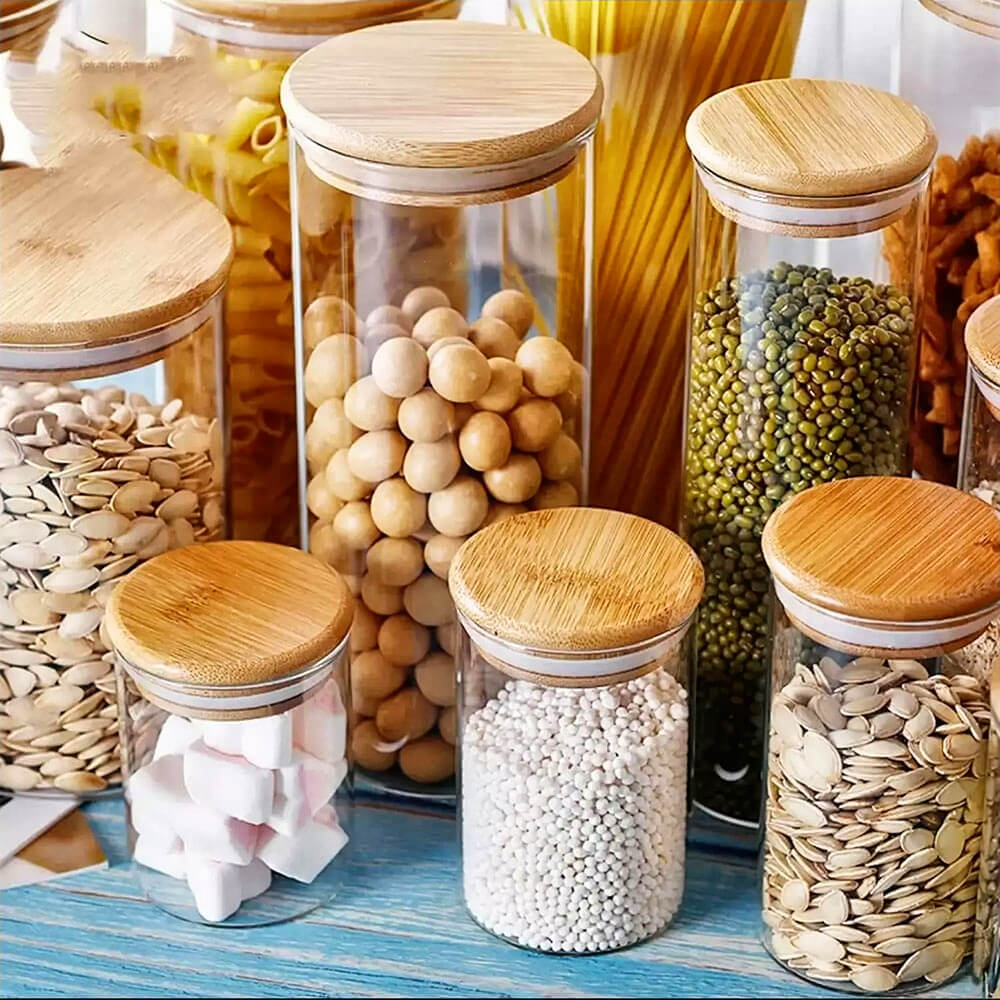 A bamboo and glass jar set displayed on a countertop, featuring multiple clear cylindrical jars filled with various dry goods like seeds, pasta, legumes, and marshmallows. Each jar has a flat bamboo lid with a silicone gasket for an airtight seal.