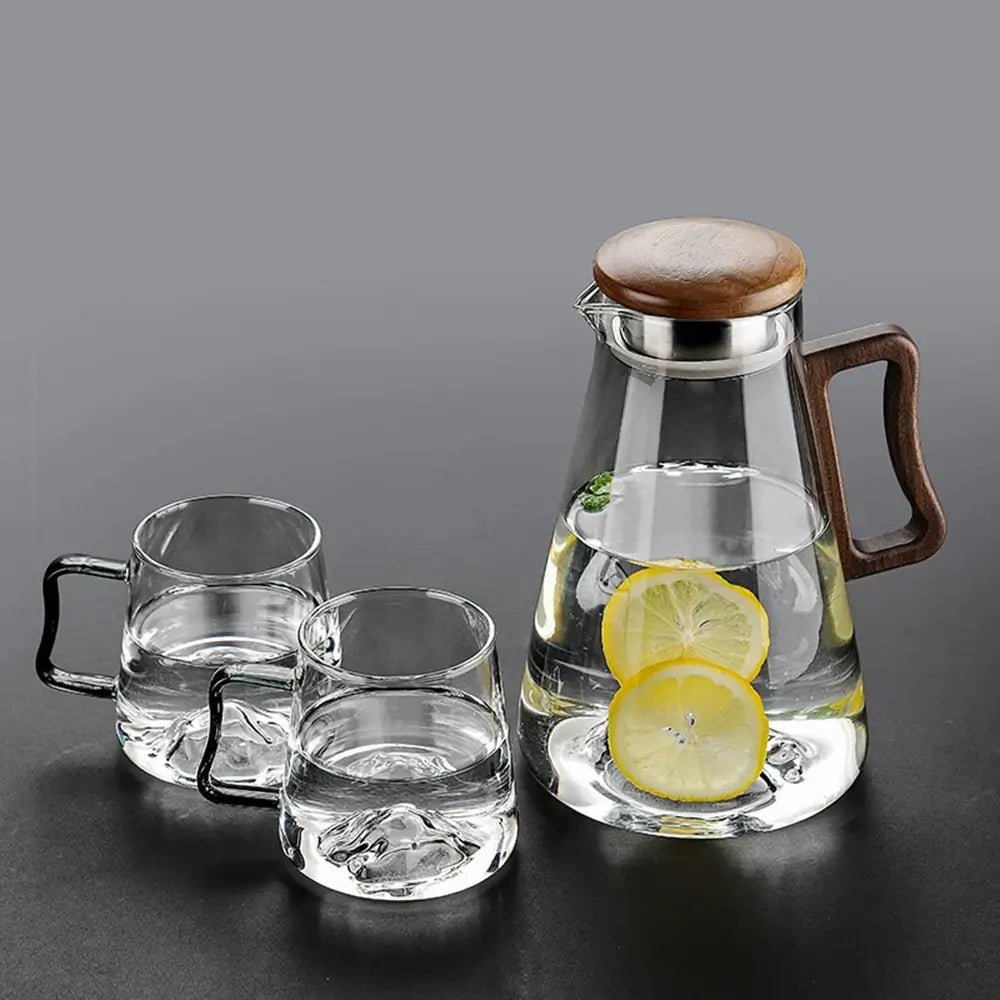 Clear glass carafe with wooden lid and handle, filled with water and lemon slices, next to two clear glass mugs on a dark surface.