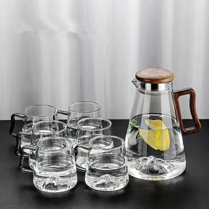 Set of glass mugs and a glass carafe with wooden lid and handle on a dark surface.