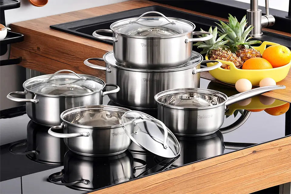 Set of stainless steel cookware on a kitchen stove with fruits and vegetables in the background.