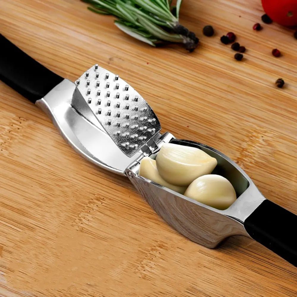 Stainless steel garlic press with black handles, holding fresh garlic cloves on a wooden surface with herbs and spices nearby.