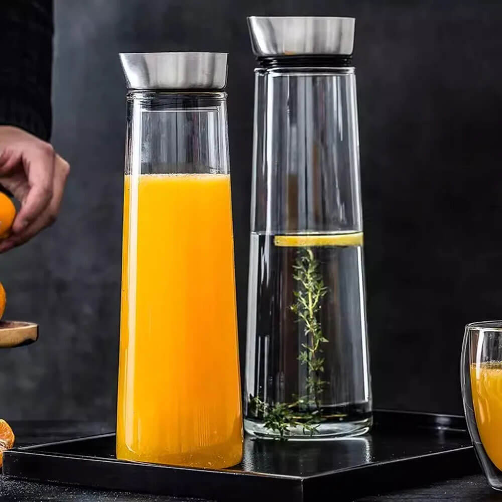 Two 1-liter glass water bottles with stainless steel leakproof lids. One contains orange juice, the other water with lemon and herbs.