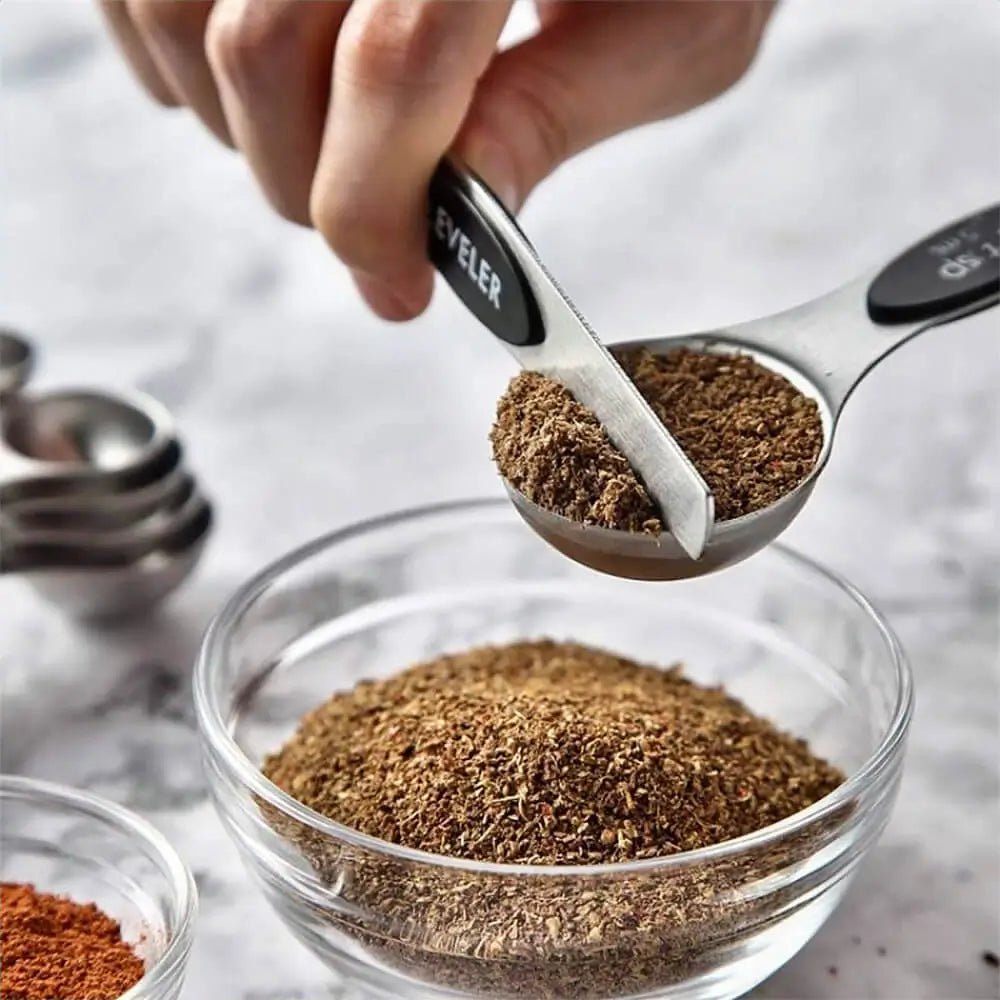 Hand using a measuring spoon to scoop spices from a glass bowl on a marble surface.