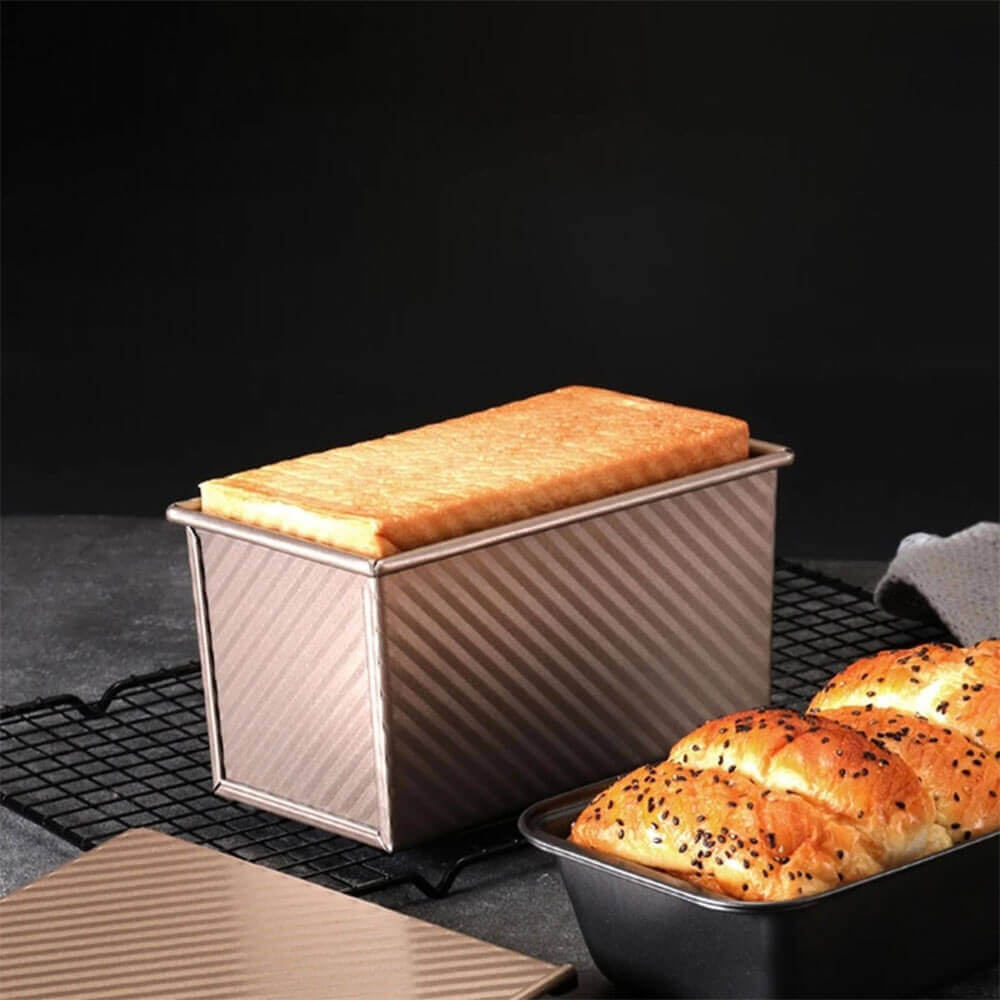 Rectangular best non stick loaf pan with baked bread inside, shown on a cooling rack beside golden brown sesame seed buns.
