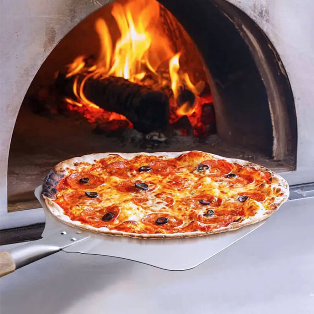 The best pizza peel with a detachable oak handle, sliding a freshly baked pepperoni pizza out of a wood-fired oven with glowing flames.