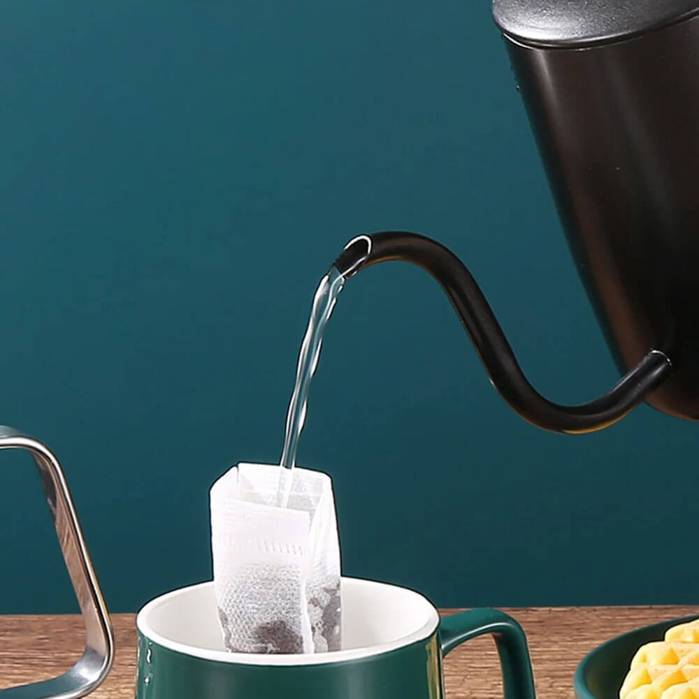 A black best pour over coffee kettle with a curved gooseneck spout pouring water into a green mug. Inside the mug, a drip coffee filter with ground coffee is visible, while the background features a teal wall and a wooden surface.