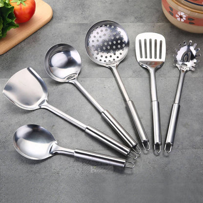 A set of six of the best stainless steel cooking utensils neatly arranged on a gray surface. The utensils include a wok spatula, ladle, slotted spoon, slotted turner, pasta server, and serving spoon, all featuring a polished stainless steel finish. The handles are smooth and ergonomically designed, with small hanging loops at the ends for convenient storage. Fresh vegetables and a ceramic pot in the background add a vibrant culinary context to the image.