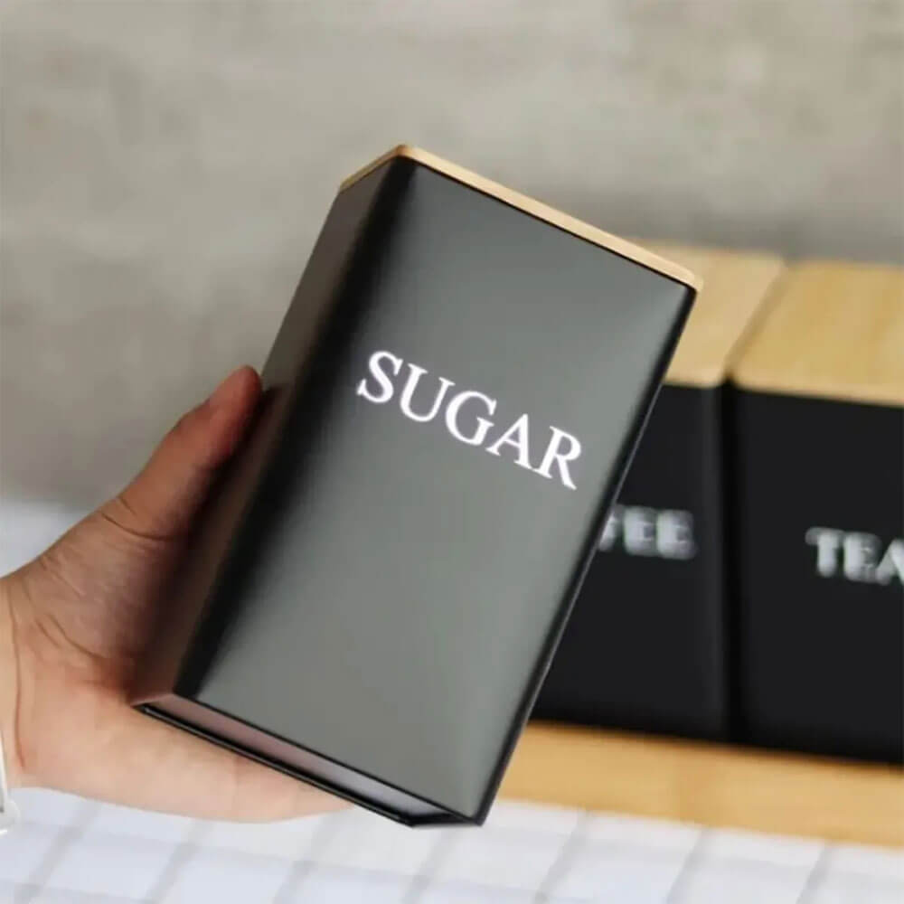 A hand holds a black stainless steel container labeled SUGAR with a wooden lid; other labeled containers are visible behind.