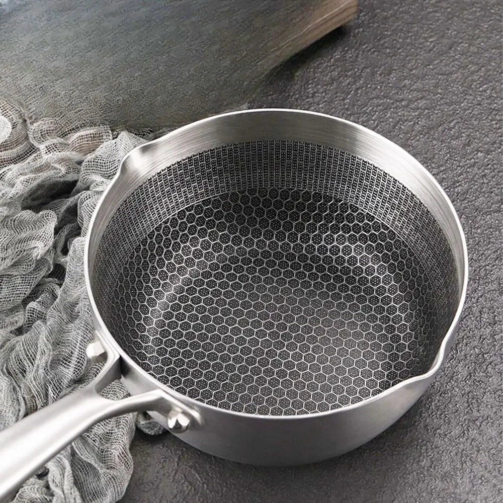 A stainless steel saucepan with a long handle and a hexagon-patterned interior, placed on a dark textured surface beside a cloth.