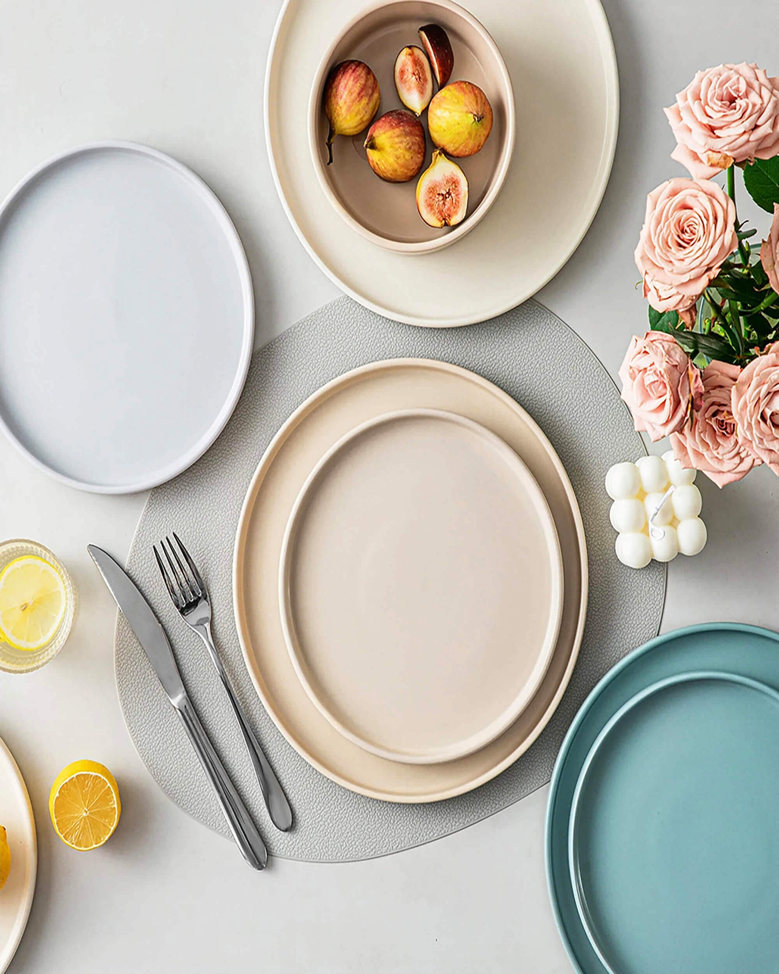 Better Homes and Gardens tableware set with neutral and pastel plates, a bowl of figs, cutlery, lemons, pink roses, and a decorative candle on a textured placemat.