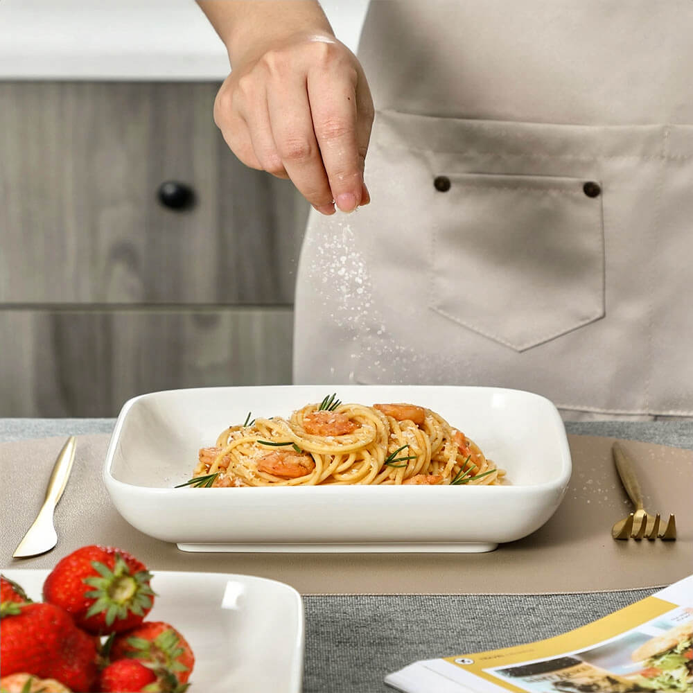  A person wearing a beige apron sprinkles seasoning over shrimp pasta served in a white porcelain big serving bowl. Another serving bowl with strawberries is placed nearby on a dining table with gold cutlery and a magazine.