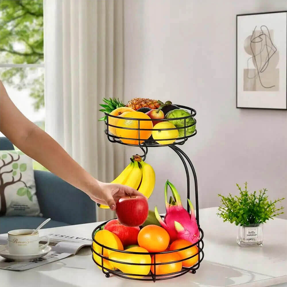 2-tier black wire fruit basket with bananas and assorted fruits and a hand is picking an apple.