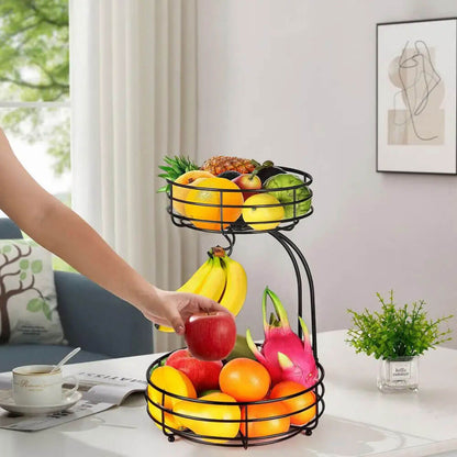 A black 2-tier wire fruit basket elegantly displayed on a countertop. The lower tier holds vibrant fruits like oranges, apples, and dragon fruit, while the upper tier features a variety of fresh produce, including a pineapple and citrus fruits. A built-in hook suspends bananas, preventing bruising and ensuring even ripening.