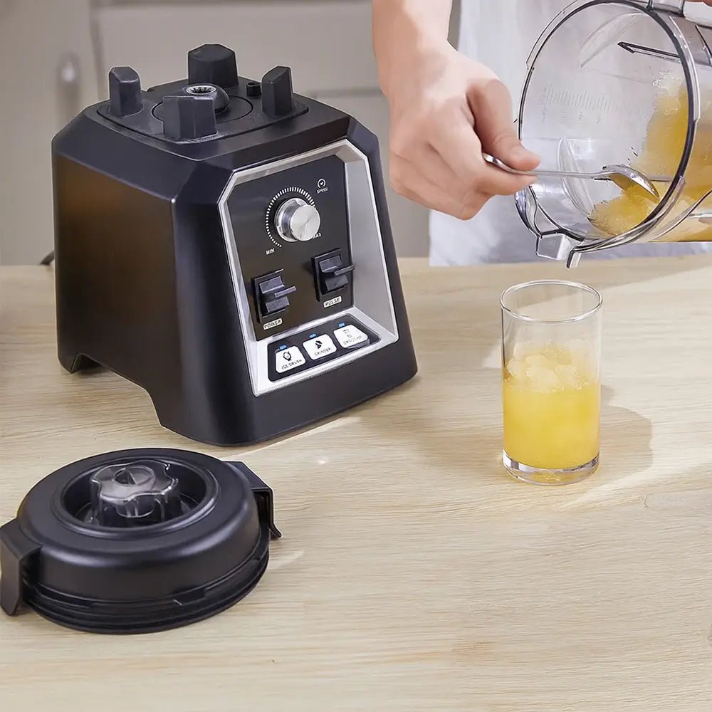 Black blender on a wooden surface with a glass of juice and a hand pouring ingredients.
