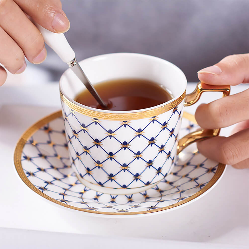 A stylish blue tea cup and saucer from the honeycomb tea set pack, featuring an elegant honeycomb pattern with golden accents. A hand is holding the cup while stirring tea with a matching spoon, highlighting the luxurious design.