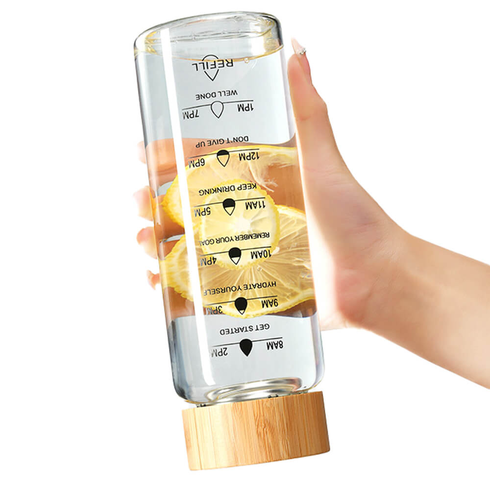 A borosilicate glass hydration bottle filled with water and lemon slices, featuring printed time markers for tracking water intake. The natural bamboo lid is shown at the bottom, adding a sustainable and stylish touch. A hand is holding the bottle, highlighting its lightweight and portable design.