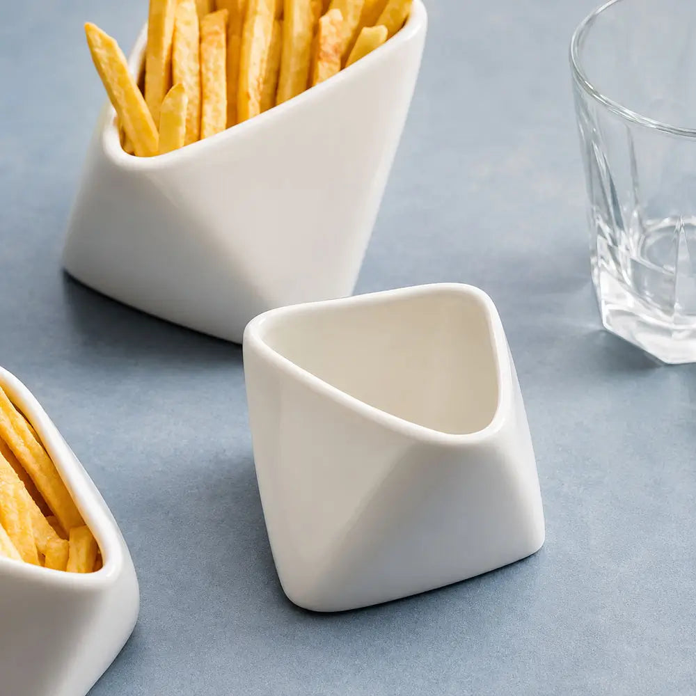 Three modern white ceramic condiment dishes with angled triangular shapes, shown on a blue-gray surface. Two dishes contain french fries, and a clear glass is partially visible on the right.