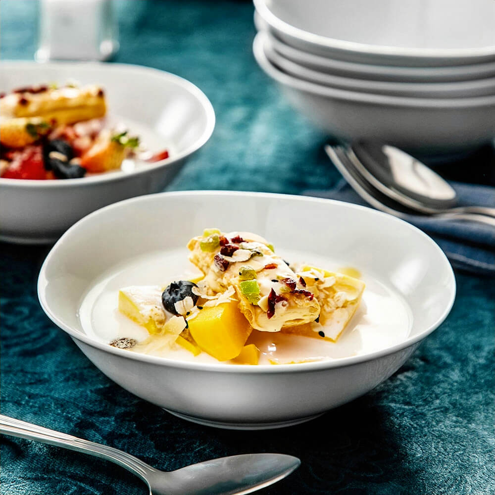 White snack bowl filled with a mix of fruits, creamy dessert, and toppings, placed on a dark tablecloth with a spoon beside it and stacked bowls in the background.