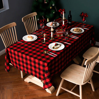 A dining table is set with a red and black buffalo plaid tablecloth, creating a warm and festive vibe. The table is adorned with plates of desserts and bread, gold utensils, and wine glasses with red napkin decorations. White candles in gold holders add an elegant touch, while a Christmas tree in the background enhances the holiday ambiance. The light wooden chairs and floor provide a cozy contrast to the bold tablecloth design.