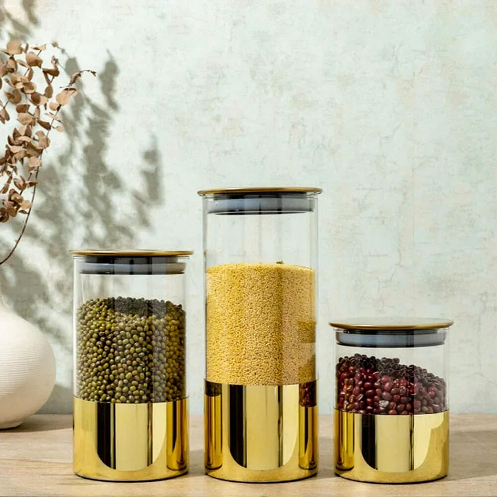 Three stackable glass canisters with gold accents are displayed on a wooden surface. Each canister is filled with dried foods, including green peas, yellow grains, and red beans, showcasing an elegant and modern design.