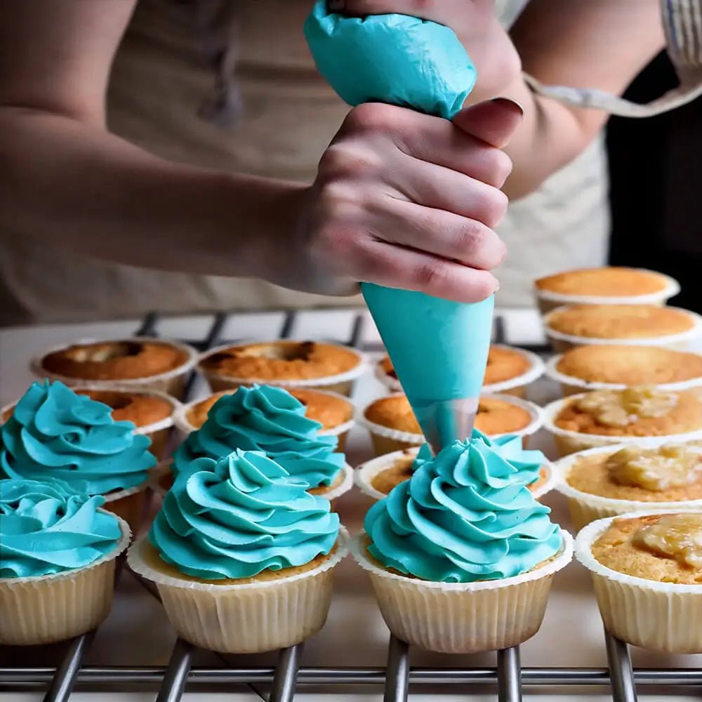 Hand using cake icing decorating set with piping bag to apply blue frosting swirls on cupcakes arranged on a cooling rack.