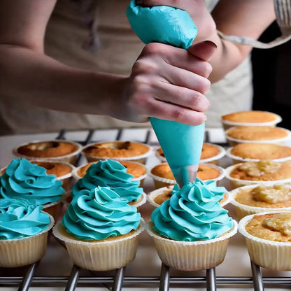 Hand using cake icing decorating set with piping bag to apply blue frosting swirls on cupcakes arranged on a cooling rack.