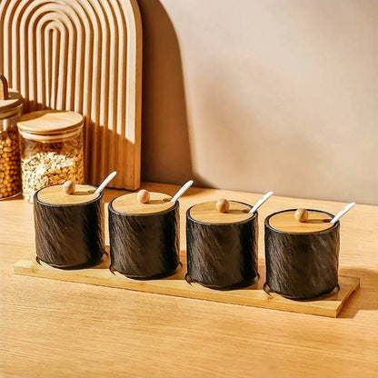 Four black ceramic jars with a textured, glossy finish sit on a rectangular bamboo tray. Each jar has a bamboo lid with a wooden knob on top, and a small white spoon rests in a slot on each lid. This elegant arrangement showcases one of the most stylish canisters sets for the kitchen, blending functionality with modern design. The setup is on a wooden surface with warm lighting, accompanied by natural decorative elements like a wooden arch and jars filled with grains in the background.