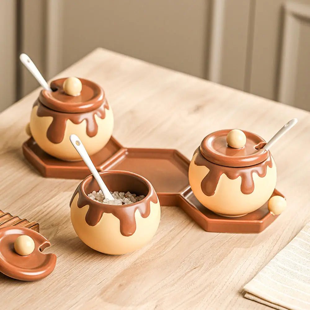 Three chocolate-drip design ceramic canisters sets for the kitchen on hexagonal trays, one open and containing small white granules.