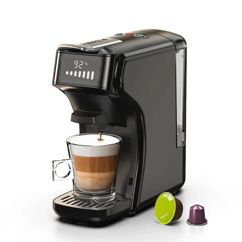 Black capsule coffee machine with digital display brewing cappuccino into glass cup, two capsules beside on white background.