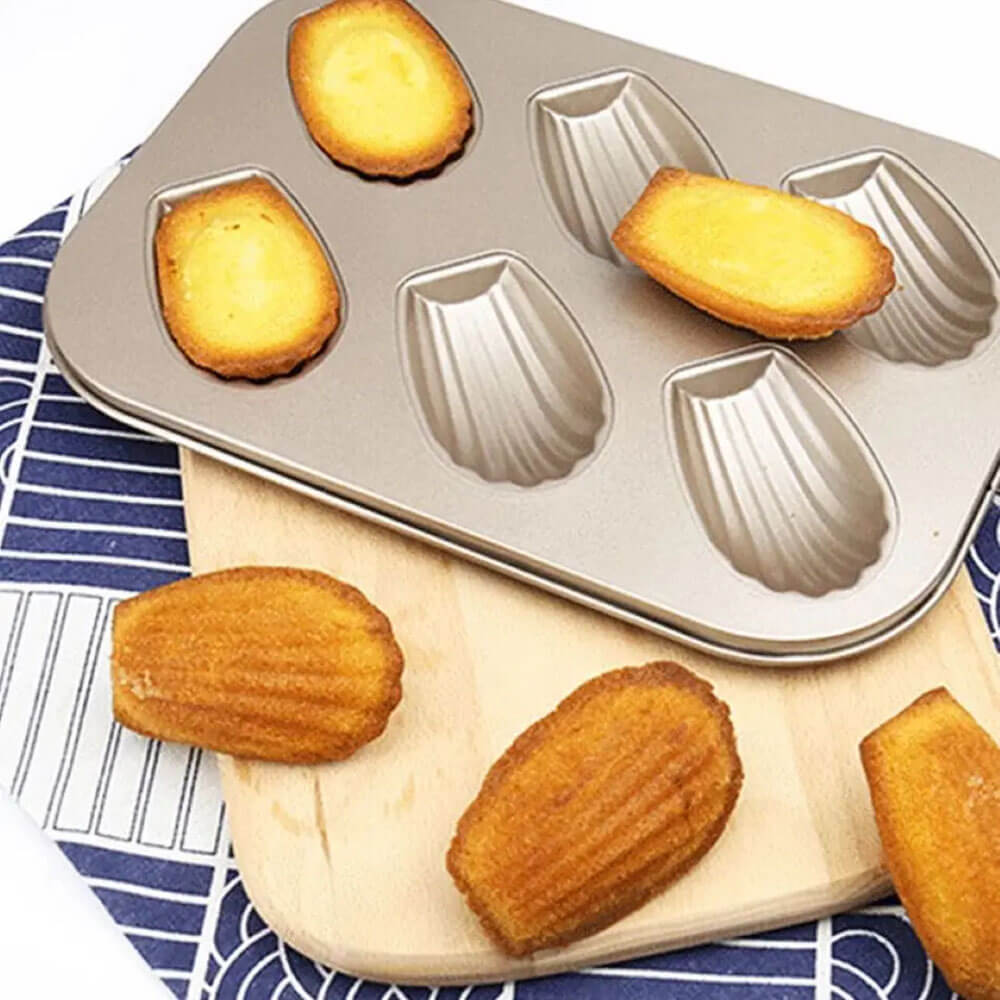 A carbon steel baking pan with six shell-shaped molds, holding a few golden-brown madeleines. Some madeleines are arranged on a wooden cutting board and a patterned fabric.