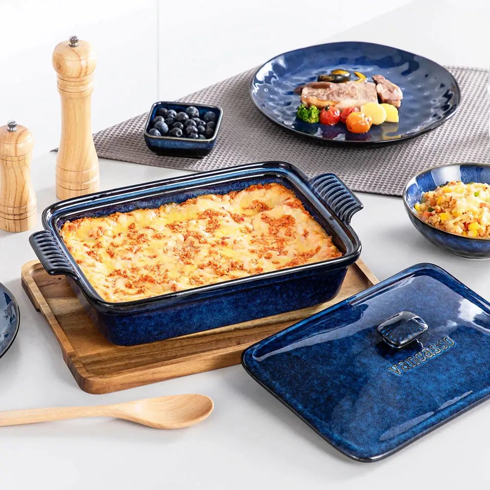 Blue porcelain casserole dish filled with baked lasagna placed on a wooden board, with its matching lid and tableware nearby.