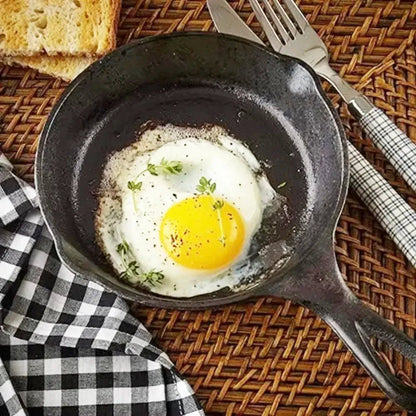 A cast iron grill skillet with a fried egg and herbs on a woven mat beside toast, fork, knife, and a checkered napkin.
