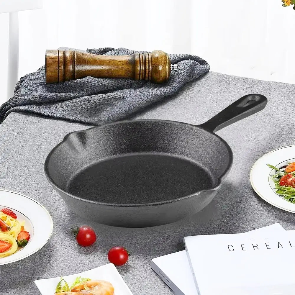 A black cast iron skillet induction pan sits on a gray tablecloth with tomatoes, plates of food, a pepper mill, and books nearby.