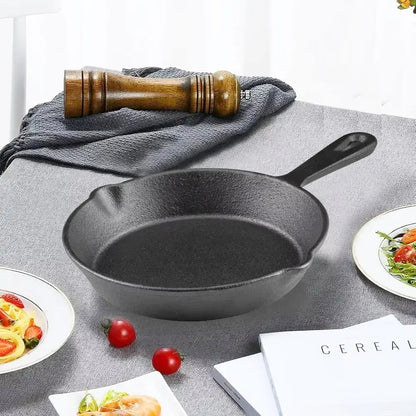 A black cast iron skillet induction pan sits on a gray tablecloth with tomatoes, plates of food, a pepper mill, and books nearby.