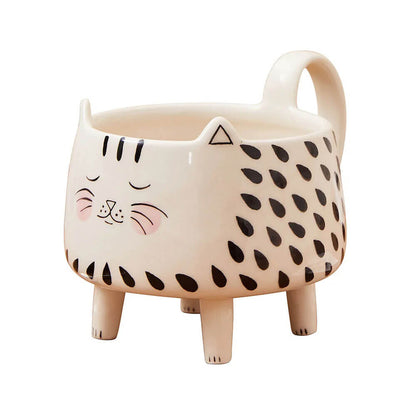 A ceramic mug shaped like a cat with a cute and playful design. The mug features a smiling cat face with closed eyes, pink cheeks, black stripes on the forehead, and black teardrop-shaped patterns on the body. It stands on four short legs and has a handle at the back.