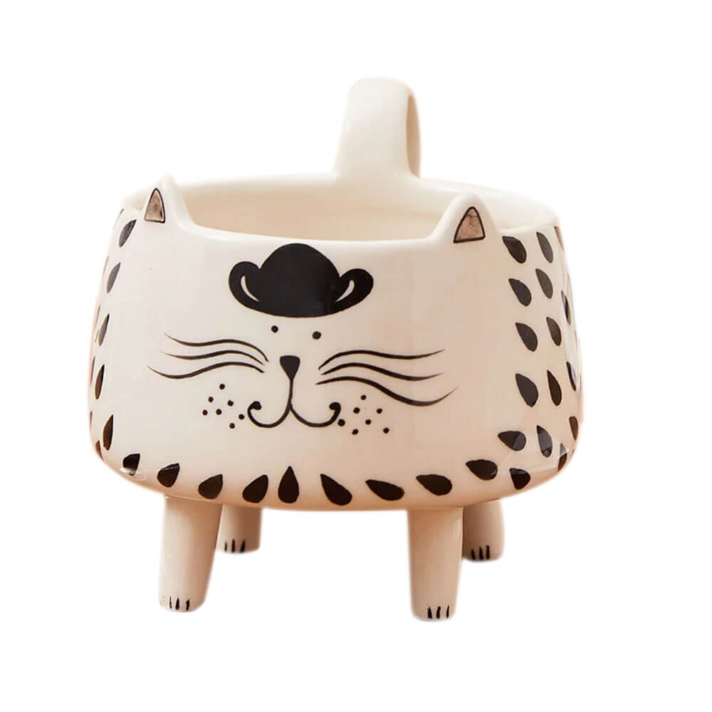 A ceramic mug shaped like a cat with a whimsical design, featuring a black cowboy hat, a smiling face with whiskers, and black teardrop-shaped patterns on its body. The mug stands on four short legs and has a handle at the back.