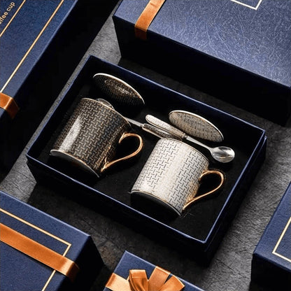 The image showcases a luxury gift set featuring two ceramic coffee mugs with lids, nestled inside a premium navy-blue box with gold accents and an elegant orange ribbon. The mugs have gold-accented handles and feature intricate geometric patterns in black and white. Each mug is paired with a matching ceramic lid and a stainless steel spoon, emphasizing both style and functionality.