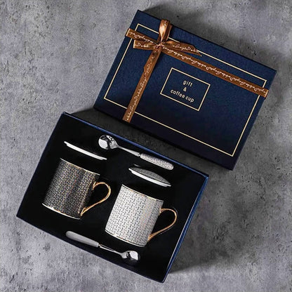 An elegant gift set featuring two ceramic coffee mugs with lids, beautifully arranged in a navy-blue box with gold detailing and a decorative ribbon. Each mug is adorned with a refined geometric pattern and gold-accented handles, accompanied by matching ceramic lids and stainless steel spoons. 