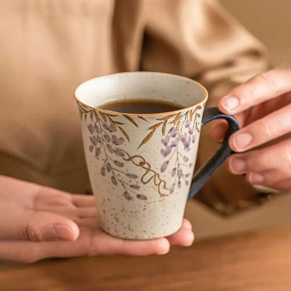 Hands holding a ceramic mug with floral pattern and dark handle, filled with coffee, resting above a wooden table.