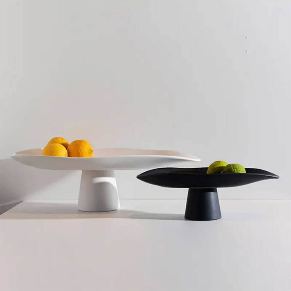 Two ceramic fruit bowls, one in white and the other in black, both with minimalist designs. The white large ceramic fruit bowl holds several oranges, while the black smaller bowl contains green limes. Each bowl features a wide, curved surface and a sturdy pedestal base, creating a modern and elegant look against the neutral background.