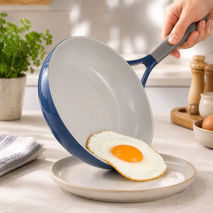 Person serving a fried egg from a blue ceramic non stick cookware set skillet onto a plate in a kitchen setting.