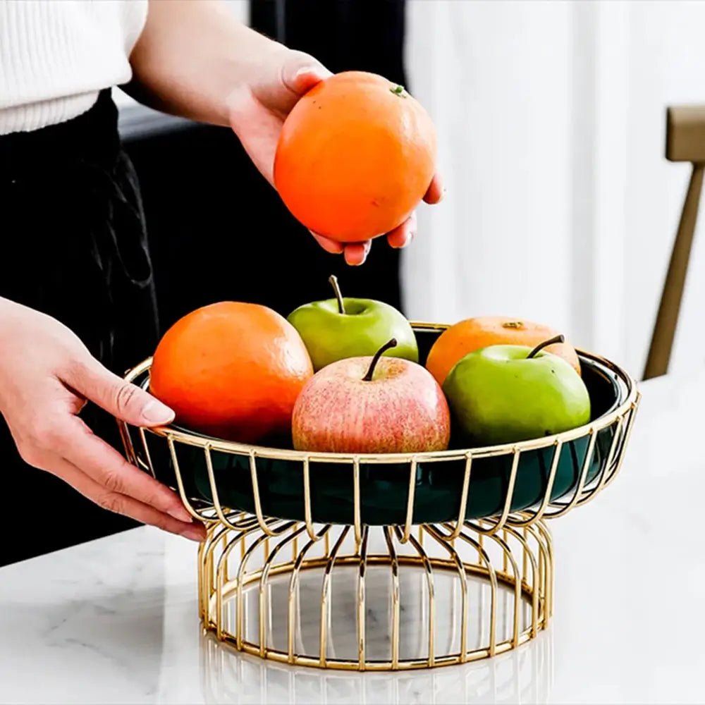 Hands placing oranges and apples into a pedestal fruit bowl with green ceramic dish and gold wire stand on kitchen counter.