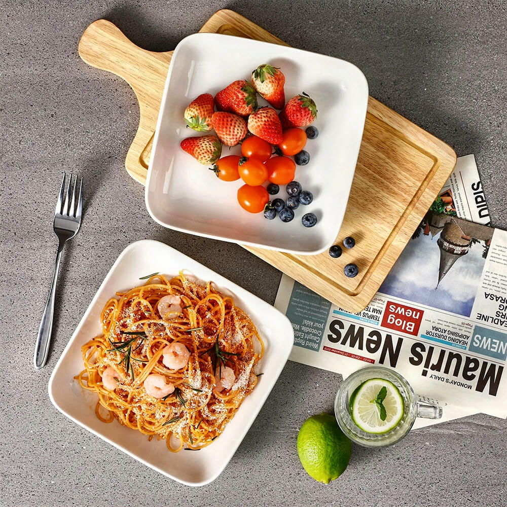 Two white ceramic serving bowls on a gray countertop, one filled with shrimp pasta and the other with strawberries, cherry tomatoes, and blueberries. A wooden cutting board, a newspaper, a fork, and a glass of lemon water with a lime are also present.