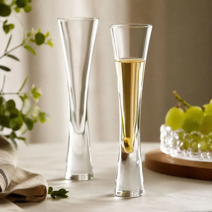 Two champagne flute glasses on a table, one filled with a sparkling drink, beside grapes, greenery and a folded cloth.