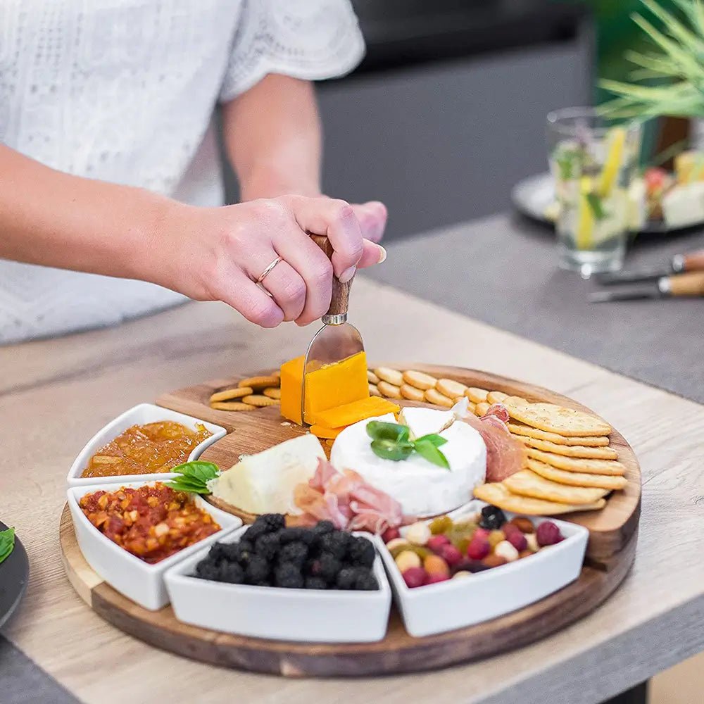 A person cuts cheddar on a wooden cheese board and knives set with crackers, brie, blackberries, nuts, and dips in ceramic bowls.