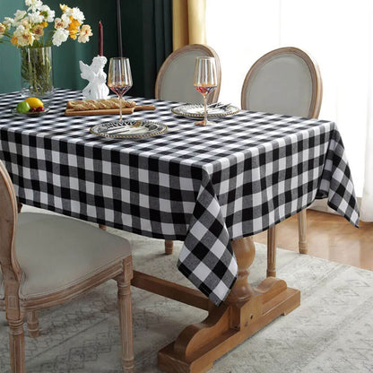 A stylish dining table is dressed with a black and white Christmas plaid tablecloth, creating a classic and festive look. The table is set with elegant plates, gold-accented wine glasses, and a centerpiece featuring a white angel figurine and a vase of fresh flowers. A wooden serving board with bread and a small fruit arrangement adds a rustic touch, while the neutral upholstered chairs and soft lighting complete the sophisticated holiday ambiance.