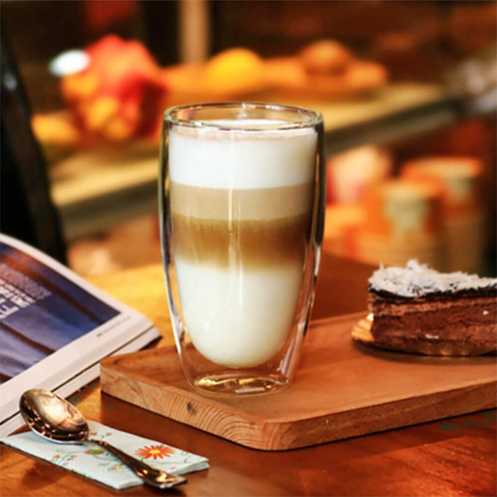 A clear glass double-walled coffee tumbler sits on a wooden tray, filled with a beautifully layered latte. Nearby, there&