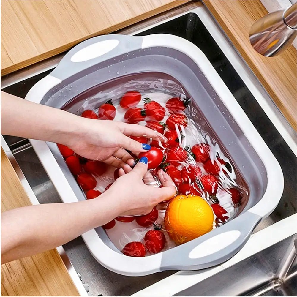 Hands washing tomatoes and an orange in a gray collapsible cutting board used as a basin inside a kitchen sink.