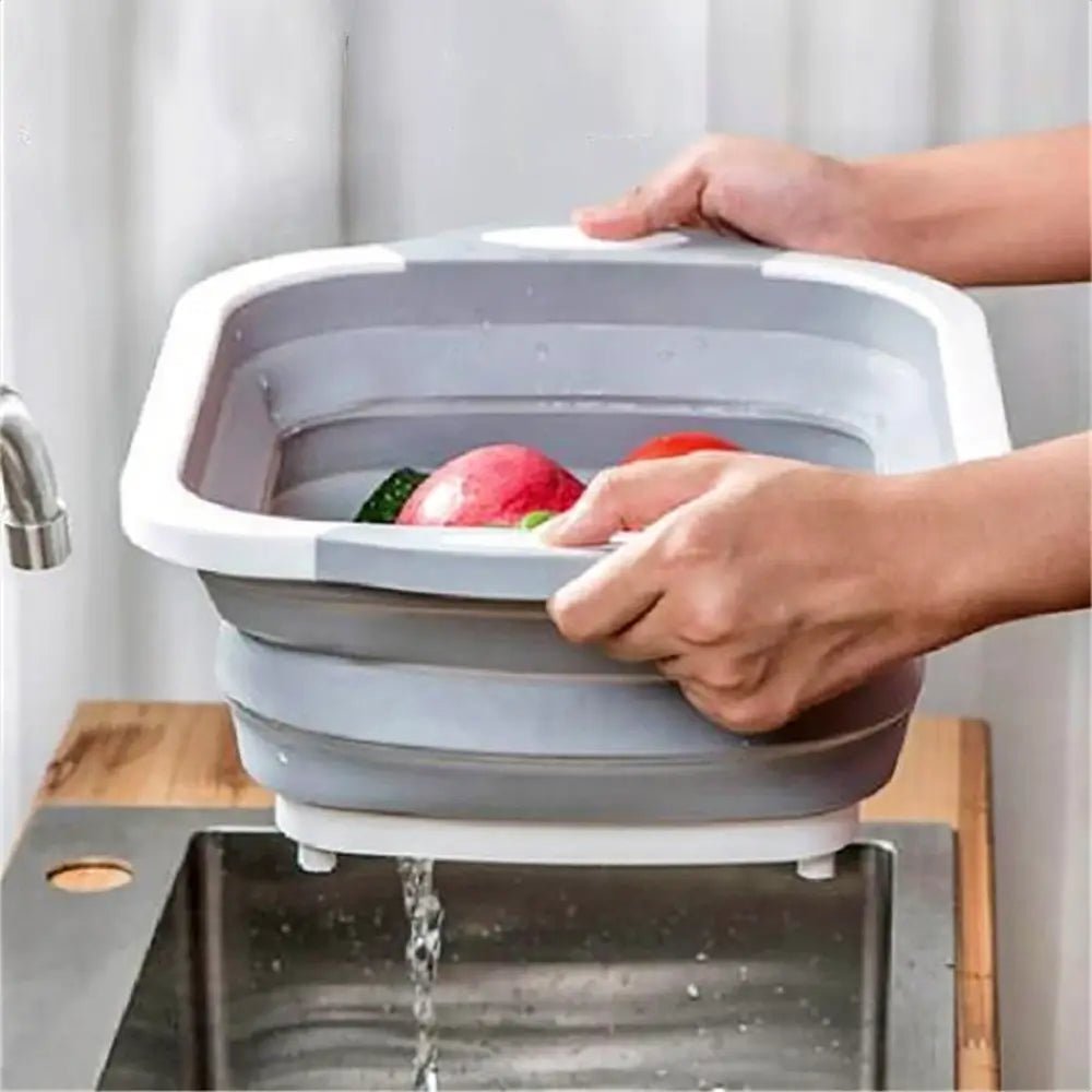 Hands draining water from a gray collapsible cutting board used as a basin, filled with rinsed fruits and vegetables over a sink.